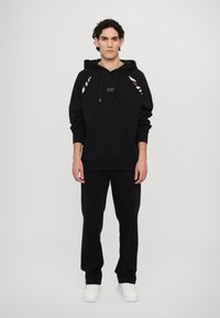 Un sweat à capuche noir avec des accents rayés, une capuche avec cordon de serrage et un patch logo, assorti à un pantalon noir et des baskets blanches. Design et coupe simples.