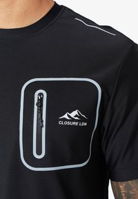 Czarny sportowy t-shirt z teksturowanej tkaniny, z szarym kieszonką na zamek błyskawiczny oraz kontrastową grafiką góry i napisem "CLOSURE LDN".
