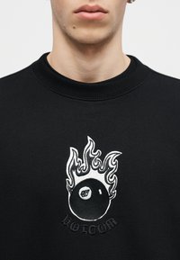 Κλείσιμο εικόνας ενός ατόμου που φοράει μαύρη φούτερ με ένα λευκό φλεγόμενο οκτάμπαλο και το κείμενο "Volcom" κεντημένο στο στήθος.