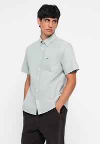 Short-sleeve button-up shirt in light grey fabric, featuring a chest pocket and small green logo embroidery. Smooth texture and tailored fit.