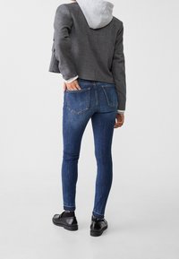 Persona vistiendo una chaqueta cortada gris sobre una sudadera gris, jeans ajustados azules y zapatos de charol negros, de pie con una mano en el bolsillo trasero.