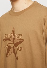 Ruskea puuvillainen pitkähiha, jossa on graafinen tähtimuotoilu ja tummemmasta ruskeasta tekstistä "Carhartt WP 313". Sujuva pinta, pyöreä kaula-aukko.