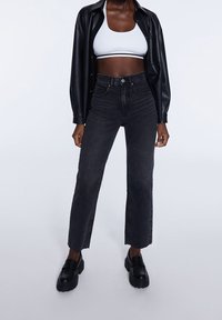 Mujer vestida con una chaqueta de cuero negra sobre una camiseta crop blanca, jeans negros de cintura alta y mocasines de plataforma negros, de pie sobre un fondo liso.
