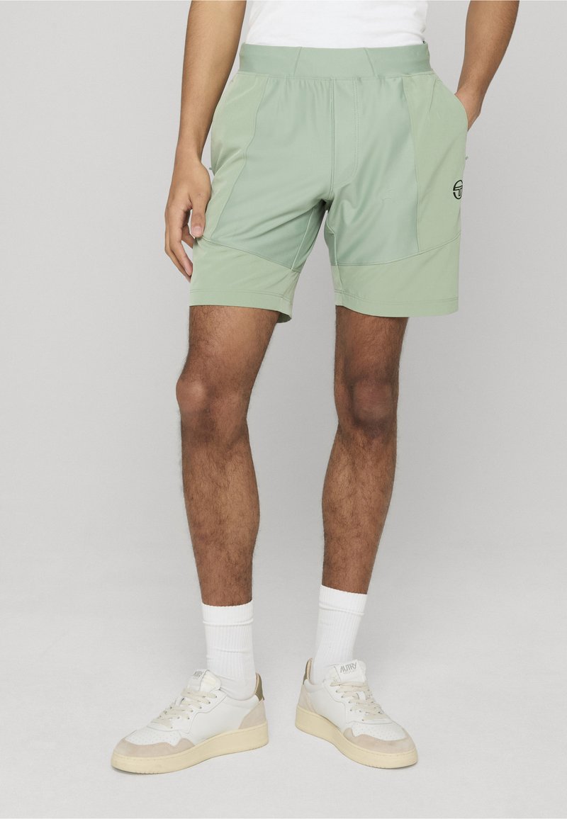 Homme portant un short de sport vert clair avec poches latérales, des chaussettes blanches et des baskets blanches, sur un fond uni.