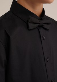 Chemise noire avec col classique et nœud papillon texturé noir, fabriquées en tissu lisse avec une fermeture à boutons.