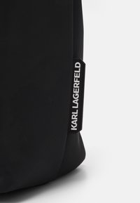 Zwarte stoffen tas met een gestructureerd oppervlak en een rechthoekig label met de tekst "KARL LAGERFELD" in het wit op een contrasterende zwarte achtergrond.