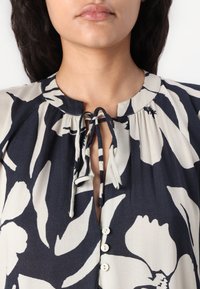 Blouse dengan pola bunga yang menampilkan dasar navy dengan bunga krim besar, leher yang diikat, dan deretan kancing di bagian depan. Kain yang lembut.