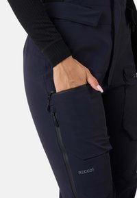 Marineblå softshellbukser med en lynlåslomme, RECCO-logo og en tætsiddende silhuet. Stoffet har en glat tekstur og en mat finish.