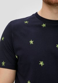 Marineblauw T-shirt met verspreide kleine groene palmboomprints, gedragen door een persoon die bovenlichaam en nek toont.