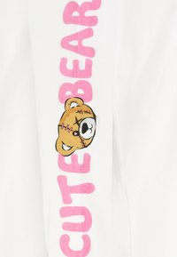 Helles Baumwollkleidungsstück mit "CUTE" und "BEAR" in Rosa bedruckt. Verfügt über eine Grafik eines genähten Teddybären mit einem lächelnden Gesicht.