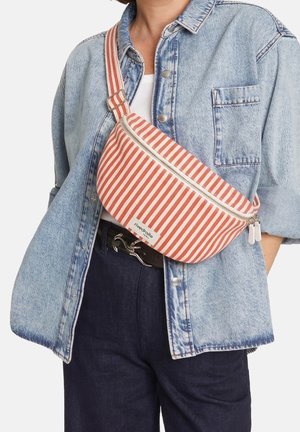 Personne portant une veste en jean claire et un pantalon foncé avec un sac bandoulière rayé rouge et blanc avec fermeture éclair, étiqueté "rivedroite."