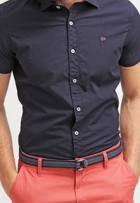 Marinblå kortärmad skjorta med knapp-down krage, med vita knappar och en liten logotyp. Kombinerad med ljus korallfärgade byxor och ett mönstrat bälte.