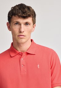 Camisa polo coral hecha de una mezcla de algodón, con cuello tipo polo, tres botones y un pequeño logotipo blanco en el pecho. Textura suave.