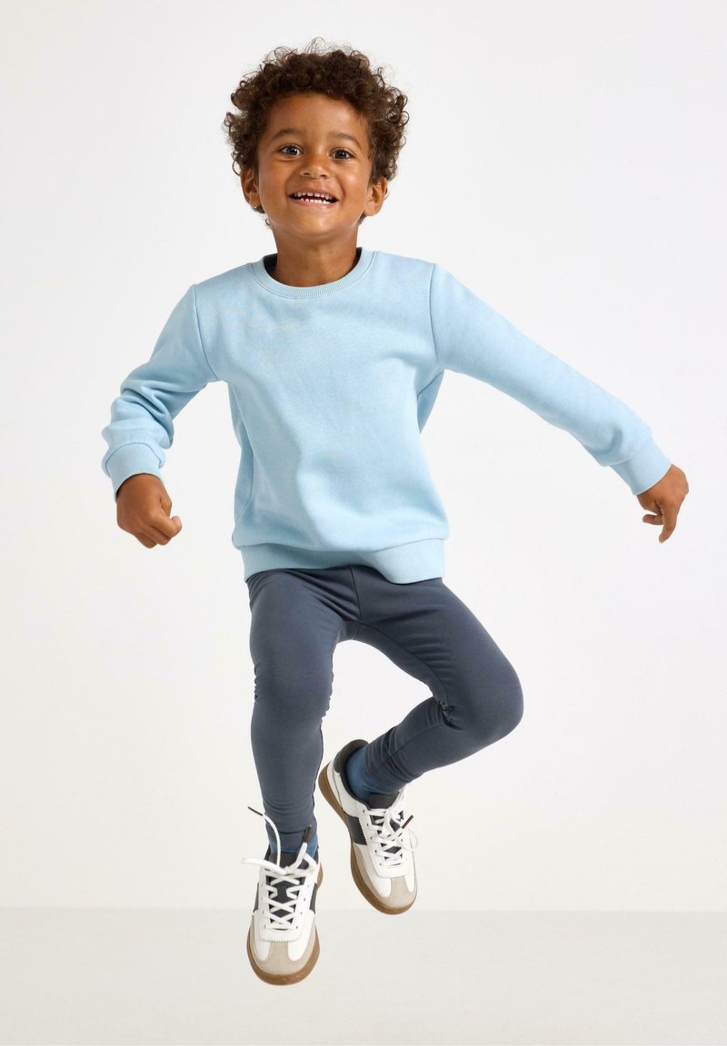 Light blue sweatshirt with ribbed cuffs, paired with dark blue leggings and white sneakers with brown accents. Child jumping against a neutral backdrop.