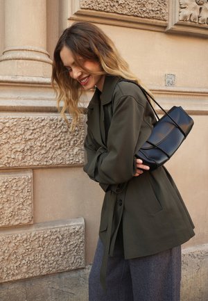 Black leather handbag with twisted cord handle held against an olive green jacket. Textured stone background adds visual contrast.