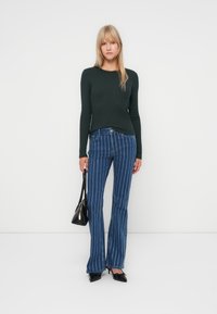 Mørkegrønn ribbet langermet topp kombinert med blå flared jeans med vertikale hvite striper, accessorert med en svart veske.