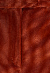 Czerwono-rdzawy materiał kordurowy z pionowymi prążkami, posiadający kieszeń boczną i gładką fakturę, co wskazuje na miękki i trwały materiał.