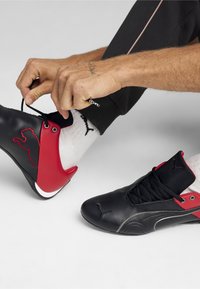 Černé a červené sportovní boty s hladkým koženým povrchem, síťovinovým jazykem a bílou podrážkou; mají výrazné logo Puma a tkaničky.