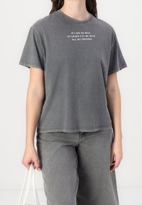 Šedé oversized tričko s kulatým výstřihem a krátkými rukávy, s bílým textem na přední straně a měkkou, opotřebovanou texturou. V kombinaci se šedými kalhotami.