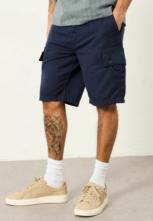 Man wearing navy cargo shorts, beige sneakers with white socks, standing with hands in pockets, showing a large tattoo on left calf.