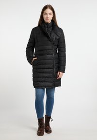 Zwarte gewatteerde jas met hoge kraag, ceintuur in de taille en horizontaal gestikt patroon. Gecombineerd met blauwe spijkerbroeken en bruine veterschoenen.