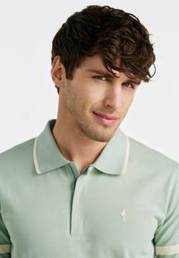 Koszulka polo w jasnozielonym kolorze z bawełny, z klasycznym kołnierzykiem i kremowymi akcentami. Posiada subtelne logo na klatce piersiowej. Miękka w dotyku.