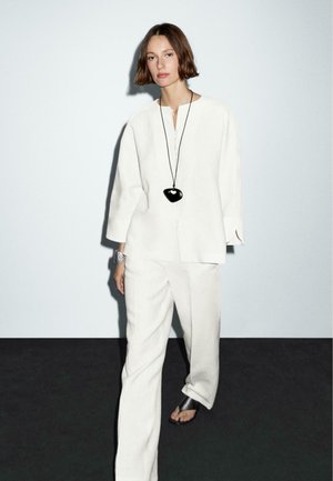 White linen outfit with a buttoned top and wide-leg trousers. Accessories include a large black pendant necklace and metallic wristwatch.