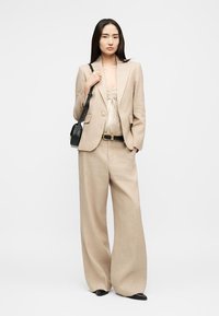 Γυναίκα που φοράει μπεζ σακάκι με ρίγες και παντελόνια φαρδιάς γραμμής, κρατώντας μαύρη τσάντα ώμου, στέκεται μπροστά σε λευκό φόντο.