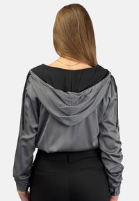 Szara kurtka z kapturem o gładkiej, błyszczącej fakturze i czarnymi wykończeniami. Posiada sznurki na kapture i długie rękawy. Widać czarne spodnie.