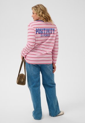 Femme aux cheveux blonds portant une chemise rayée rose et rouge avec le texte "Positivity is Power", jean bleu, tenant un sac marron, debout de dos.