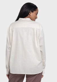 Light beige long-sleeve shirt with a collared neck, soft texture, and buttoned cuffs, worn backward, revealing simple stitched detailing.