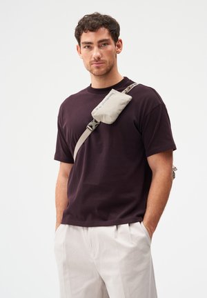 Mann trägt ein dunkelviolettes T-Shirt und hellbeige Hosen mit einer kleinen beigen Umhängetasche, die quer über seine Brust getragen wird.