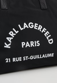 Sac noir avec texte blanc indiquant « Karl Lagerfeld Paris » en arc au-dessus de « 21 Rue St-Guillaume » sur une surface de tissu lisse.