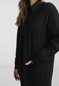 Svart stickad cardigan med avslappnad passform, långa ärmar, V-ringning och två framfickor. Texturerad tyg ger djup.