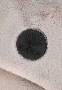 Tejido de pelaje gris con una textura suave, que presenta un logo circular de metal negro que dice "JOOP! JEANS" y un diseño de emblema floral.