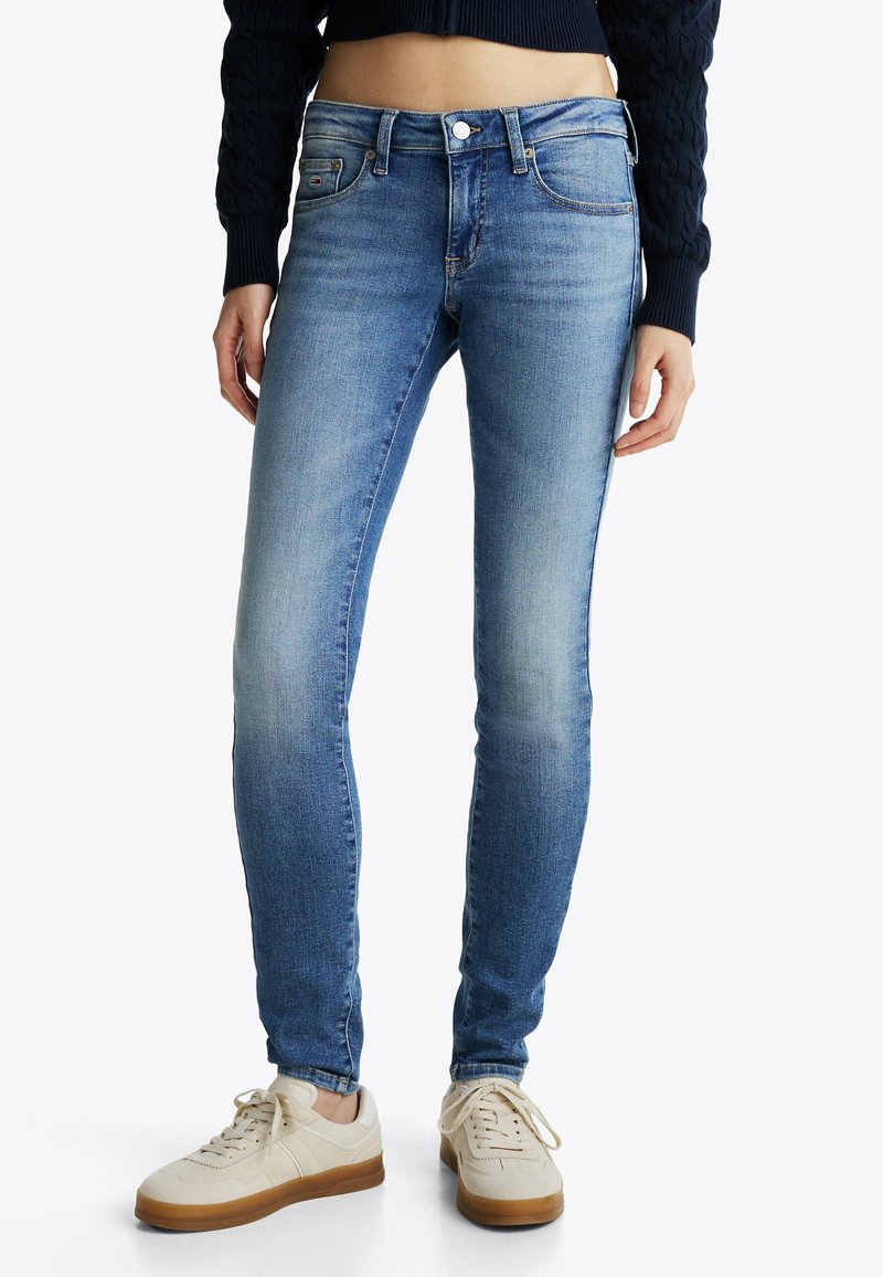 Jeans ajustados de color azul con una cintura de altura media, con un ligero desgaste y cinco bolsillos. Combinados con zapatillas crema que tienen una suela de goma.