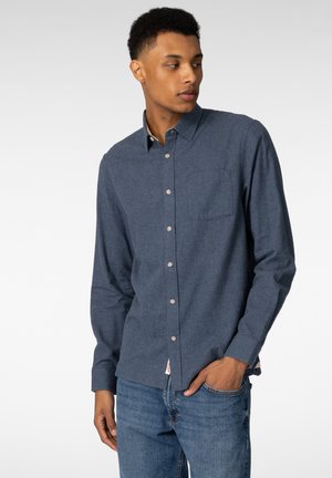 Ung mann med kort krøllete hår som har på seg en knappet mørkeblå langermet skjorte og blå jeans, med hånden i lommen, ser ut til siden.