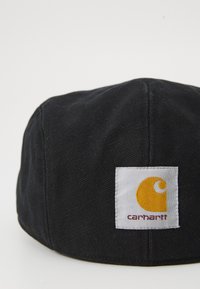 Cappello di tessuto nero con una parte superiore arrotondata e cuciture. Presenta un'etichetta quadrata bianca con un logo giallo e il testo "Carhartt".