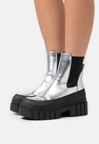 Botas de tobillo metálicas plateadas con inserciones elásticas en los laterales, suelas de goma negra gruesa y una textura suave. Cuentan con una punta redondeada y una forma única.