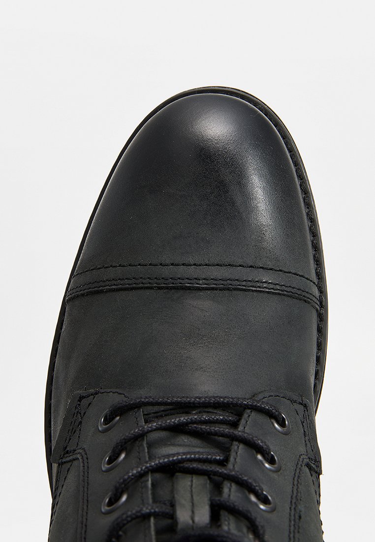 Zapato de cuero negro con punta redondeada, cierre de cordones y detalles visibles de costura a lo largo de la parte superior y la suela. Textura lisa.