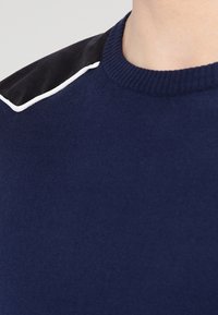 Maglione blu navy con scollatura a coste, dettagli in velluto nero sulle spalle e bordino bianco lungo la cucitura delle spalle. Tessuto liscio.