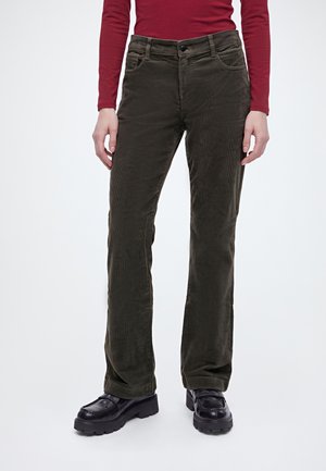 Modèle portant un pantalon en velours côtelé vert olive foncé à jambes droites, un haut bordeaux et des mocassins noirs à semelles épaisses, sur un fond blanc uni.