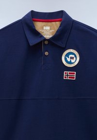 Marineblå poloshirt med to træknapper, et rundt mærke med kort og flag samt et rektangulært flagmærke på brystet.