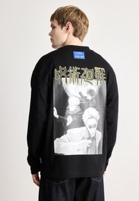 Μαύρο πουκάμισο με μακριά μανίκια που διαθέτει μια μεγάλη μονόχρωμη γραφική παράσταση στην πλάτη με στιλιζαρισμένους χαρακτήρες και κείμενο. Περιλαμβάνει μια μπλε ετικέτα μάρκας στον ώμο.