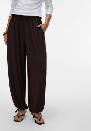 Žena v voľných čiernych nohaviciach so zúženými manžetami, v čiernych sandáloch na podpätku a bielom tričku s dlhým rukávom, s rukou v vrecku.