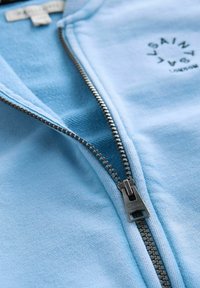 Sudadera de cremallera azul claro con cremallera metálica, cuello acanalado y un logo circular negro con texto en la zona del pecho izquierdo.