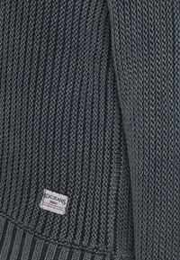 Tissu tricoté côtelé gris foncé avec une petite étiquette rectangulaire blanche BDG JEANS cousue près du bord inférieur.