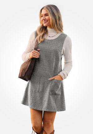 Jeune femme portant une robe sans manches à carreaux par-dessus un haut blanc à manches longues, tenant un sac à bandoulière marron, souriante et regardant sur le côté.