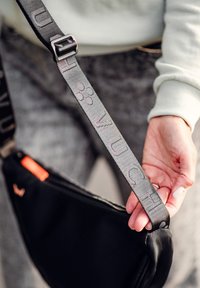 Černá ramenní taška s texturovaným popruhem, který nese embosovaný nápis. Popruh obsahuje kovovou sponu a malou oranžovou visačku.