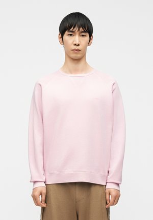 Young man with short black hair wears a light pink sweatshirt and brown pants, standing against a plain white background.